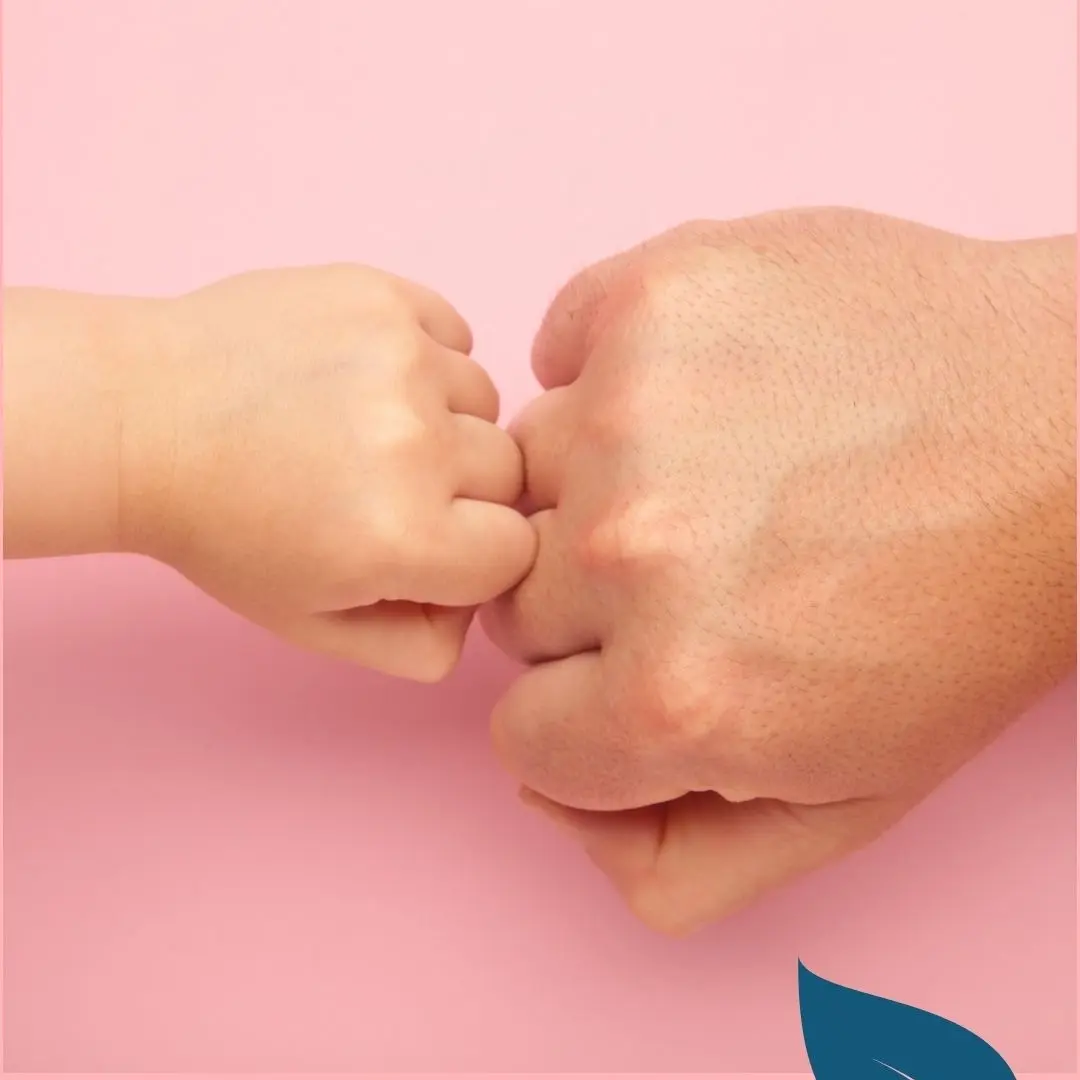Mano de un niño y de un adulto tocándose los puños sobre fondo rosa, simbolizando confianza y vínculo seguro.