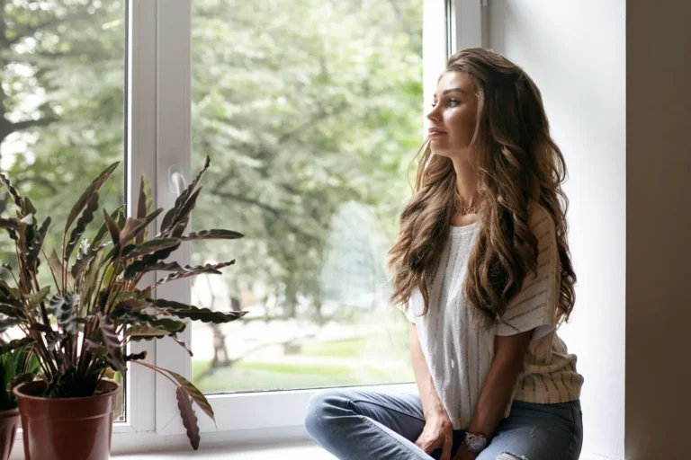 Mujer reflexiva junto a la ventana, cuidando su bienestar emocional