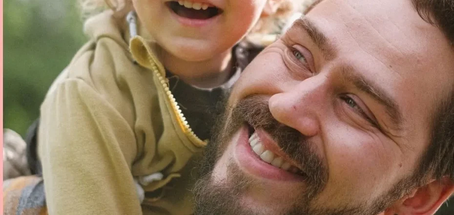Padre sonriente con su hijo pequeño sobre los hombros, simbolizando vínculo afectivo y crianza respetuosa.